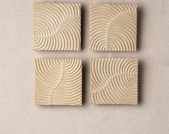 May include: Four square, beige coasters with a carved, concentric semi-circle design. The coasters are arranged in a square formation, each displaying a unique pattern of curved lines. The neutral colour and textured surface suggest a natural, earthy aesthetic.