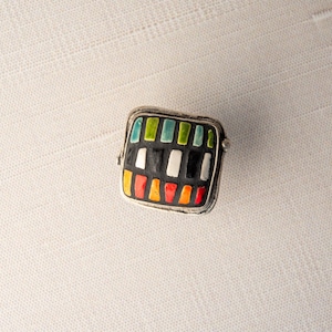 A silver ring with a square shaped ceramic tile top. The tile is decorated with a colorful tile in shades of red, orange, green, blue, black and white. Handmade in USA by ceramic and mosaic artists Romy and Clare, Jasper, IN.