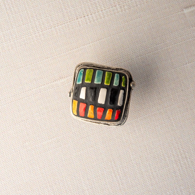 A silver ring with a square shaped ceramic tile top. The tile is decorated with a colorful tile in shades of red, orange, green, blue, black and white. Handmade in USA by ceramic and mosaic artists Romy and Clare, Jasper, IN.