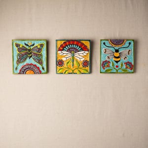 Pode incluir: Três azulejos de cerâmica com desenhos florais coloridos e motivos de insetos. O primeiro azulejo apresenta uma borboleta com asas azuis e verdes, o segundo azulejo apresenta uma libélula com asas vermelhas e amarelas, e o terceiro azulejo apresenta uma abelha com listras pretas e amarelas.