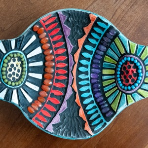 May include: A colorful mosaic dish with a fish shape. The dish is made of small, colorful tiles and has a black background. The dish is decorated with geometric patterns and has a turquoise border.