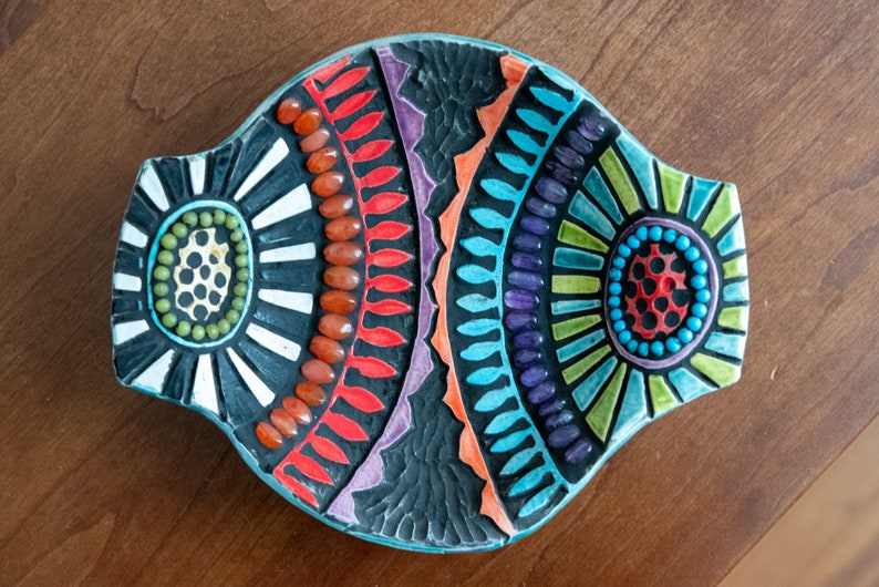 May include: A colorful mosaic dish with a fish shape. The dish is made of small, colorful tiles and has a black background. The dish is decorated with geometric patterns and has a turquoise border.