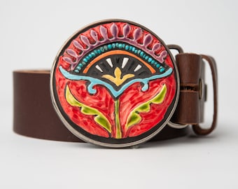 May include: A brown leather belt with a round, colourful, enamel buckle featuring a stylised floral design. The buckle has a black background with red, green, yellow, blue and purple accents.