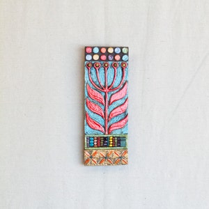 May include: A colorful ceramic tile with a stylized red flower design on a blue background. The tile is decorated with colorful beads and dots.