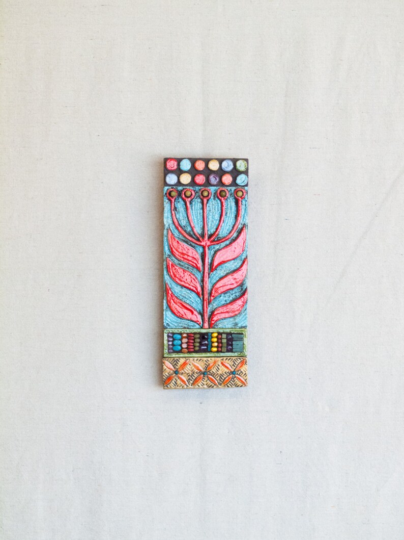 May include: A colorful ceramic tile with a stylized red flower design on a blue background. The tile is decorated with colorful beads and dots.