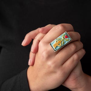 以下が含まれることがあります： 赤色の中心を持つ様式化された花を特徴とする、長方形の青、黄、緑のエナメルデザインの銀の指輪です。