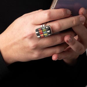 May include: A silver ring with a black and white rectangular setting. The setting is filled with colorful mosaic tiles in shades of red, orange, yellow, green, blue, and white.