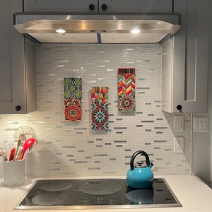 May include: Three colorful tile wall art pieces with geometric patterns in shades of red, blue, green, yellow, and white. The art is hung on a white subway tile backsplash in a kitchen with a stainless steel range hood and a blue kettle on the counter.