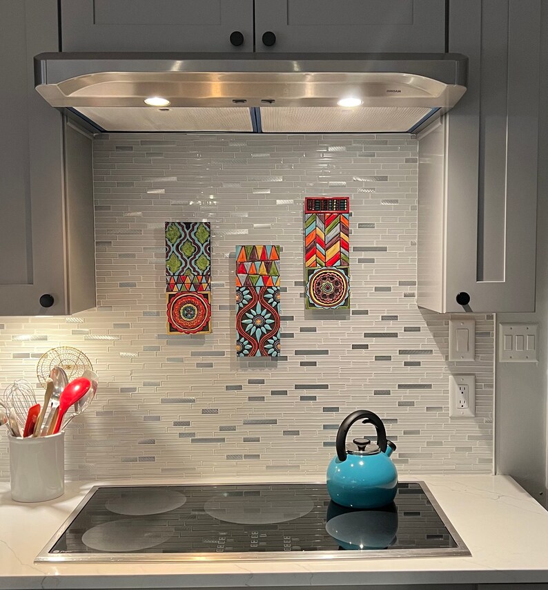 May include: Three colorful tile wall art pieces with geometric patterns in shades of red, blue, green, yellow, and white. The art is hung on a white subway tile backsplash in a kitchen with a stainless steel range hood and a blue kettle on the counter.