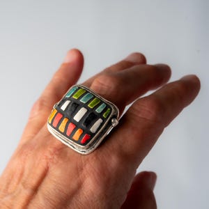 A silver ring with a square shaped ceramic tile top shown on a the third finger of a woman's left hand. The tile is decorated with a colorful tile in shades of red, orange, green, blue, black and white. Handmade in USA by ceramic and mosaic artists Romy and Clare, Jasper, IN.