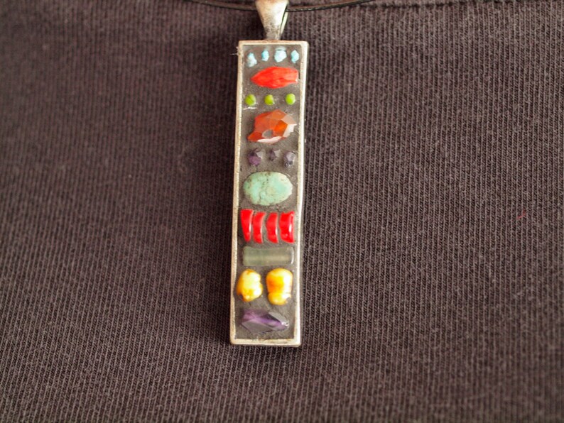 May include: A silver pendant necklace with a rectangular charm featuring a mosaic of colorful stones, including red, orange, yellow, green, blue, and purple.
