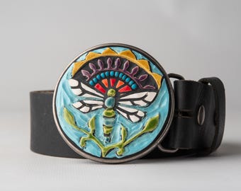May include: A black leather belt with a round, colourful ceramic buckle featuring a dragonfly design. The buckle has a blue background with a white dragonfly, green leaves, and yellow, red, and purple accents.