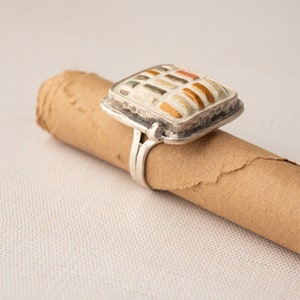May include: A silver ring with a square-shaped stone set in the center. The stone is made of small, colorful tiles arranged in a geometric pattern.