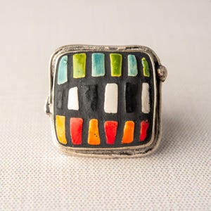 A silver ring with a square shaped ceramic tile top. The tile is decorated with a colorful tile in shades of red, orange, green, blue, black and white. Handmade in USA by ceramic and mosaic artists Romy and Clare, Jasper, IN.