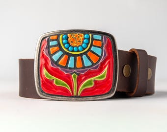 May include: A brown leather belt featuring a rectangular metal buckle with a colourful enamel inlay. The design displays a stylised flower motif in vibrant shades of red, orange, light blue, and green, accented by small turquoise beads.