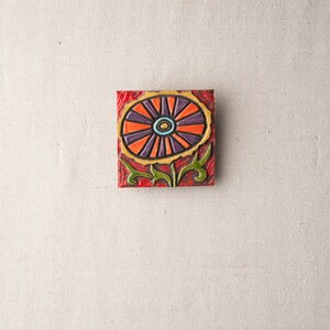 May include: A square ceramic tile with a colorful floral design. The flower is orange, purple, and yellow with a green stem and leaves. The tile has a red background.