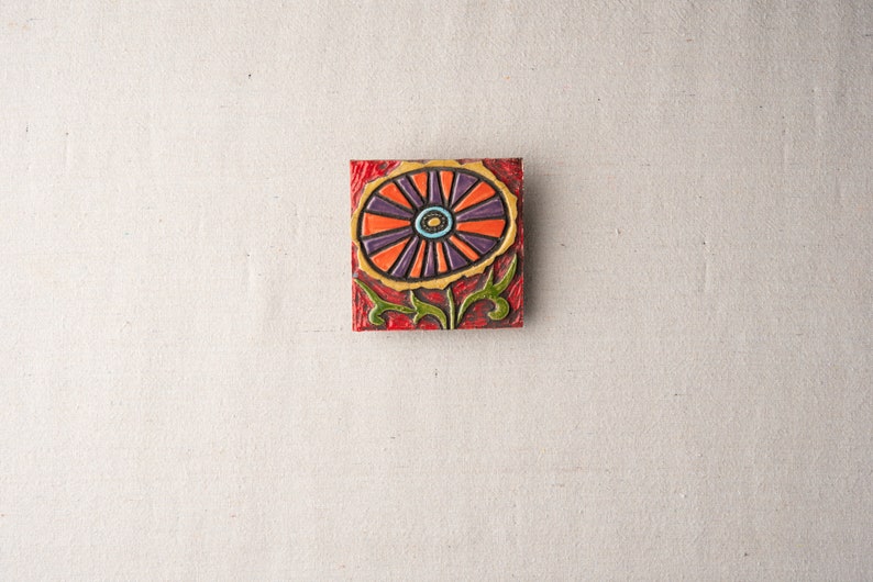 May include: A square ceramic tile with a colorful floral design. The flower is orange, purple, and yellow with a green stem and leaves. The tile has a red background.