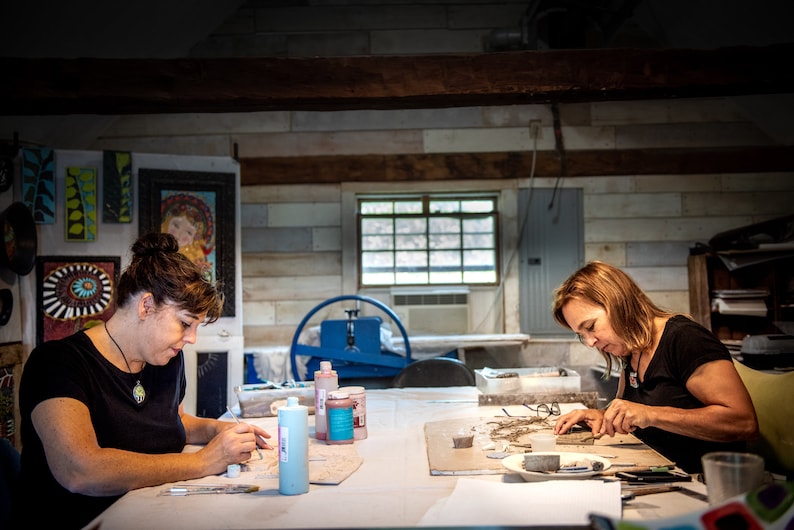 May include: Two people are working on mosaic art projects at a table. They are using small pieces of tile and glass to create their designs. There are bottles of glue and other supplies on the table.
