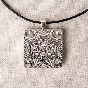 May include: A silver pendant with a circular pattern on a black cord necklace. The pendant is square shaped and has a textured surface.