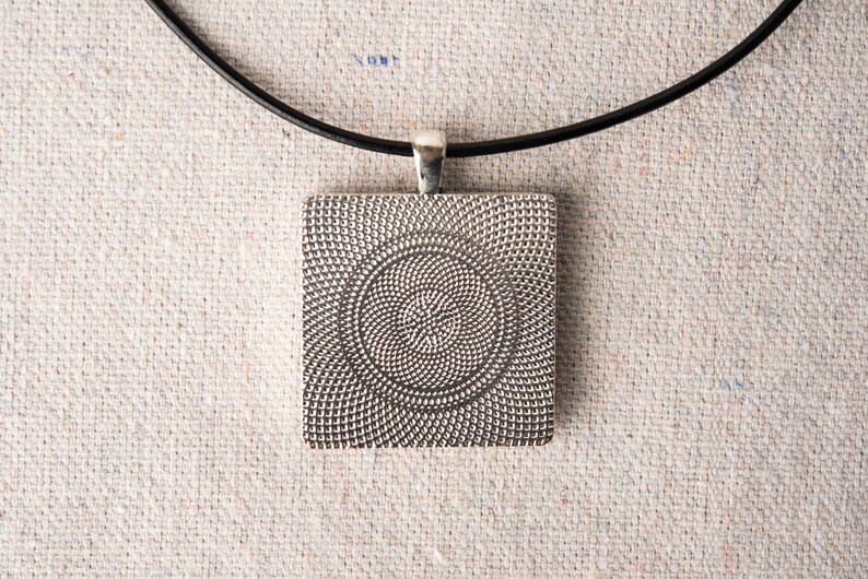 May include: A silver pendant with a circular pattern on a black cord necklace. The pendant is square shaped and has a textured surface.