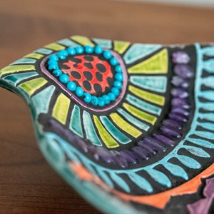 May include: A colorful ceramic dish with a geometric design. The dish is decorated with turquoise, green, purple, orange, and black paint. The center of the dish features a circle of turquoise beads.