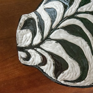 May include: A handcrafted ceramic leaf-shaped dish with a textured surface. The dish features a contrasting design of white and black, with a glossy finish. The leaf design is carved into the surface, creating a unique decorative piece.