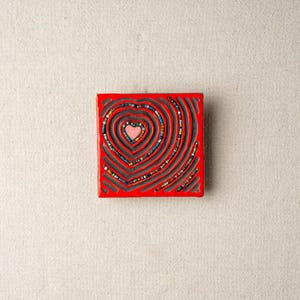 May include: A red square tile with a heart design made of colorful beads. The heart is surrounded by concentric circles of beads.