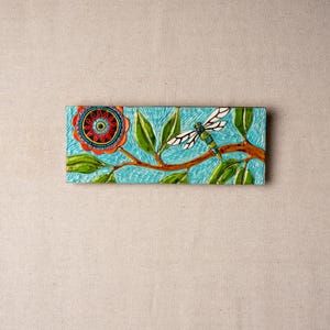 May include: A ceramic tile with a blue background featuring a stylized flower with red, orange, yellow, and green accents. A dragonfly with white wings and a green body is perched on a branch.
