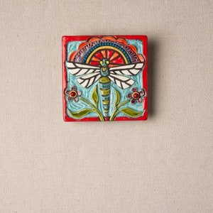 Könnte beinhalten: Eine farbenfrohe Keramikfliese mit einem Libellen-Design. Die Libelle ist blau, grün und weiß mit roten Akzenten. Die Fliese ist von einem roten Rand umgeben und hat ein florales Design.
