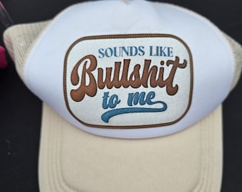 May include: A beige and white trucker hat with the phrase "SOUNDS LIKE Bullshit to me" in brown and blue lettering. The hat has a mesh back and a curved brim.