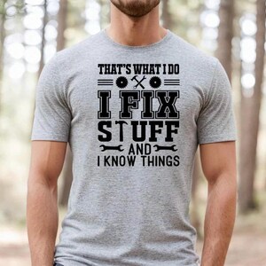 Könnte beinhalten: Ein hellgraues T-Shirt mit dem Text "THAT'S WHAT I DO I FIX STUFF AND I KNOW THINGS" in Schwarz. Das Design beinhaltet Werkzeuge wie eine Säge, einen Schraubenschlüssel und einen Hammer. Das Shirt ist kurzärmelig.