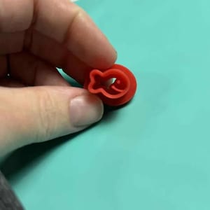 May include: A small, red, fish-shaped cookie cutter. The cutter has a circular base with a fish silhouette cut out in the center. The cutter is held by a hand against a teal background.