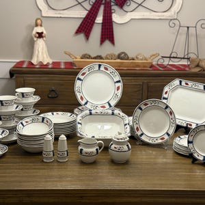 May include: A complete white dinnerware set with a blue and red floral design. The set includes plates, bowls, cups, saucers, a serving platter, a creamer, a sugar bowl, and salt and pepper shakers. The dishes are arranged on a wooden table.