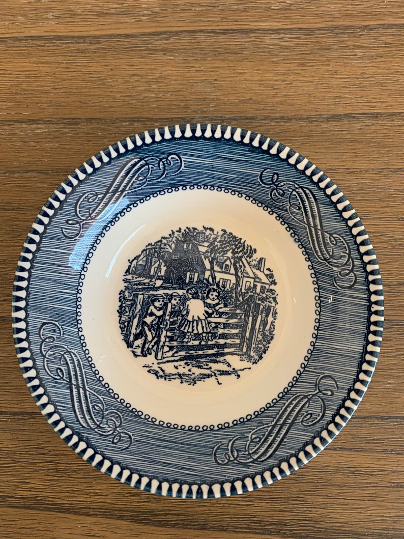 May include: A blue and white ceramic saucer with a decorative border and a black and white illustration of children playing near a house.