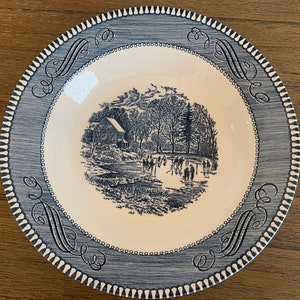 May include: A blue and white ceramic plate with a winter scene of people ice skating in front of a farmhouse. The plate has a blue and white border with a decorative pattern.