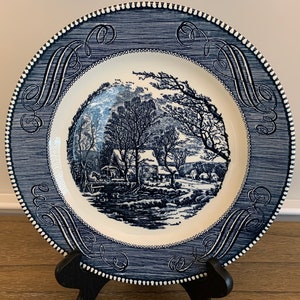 May include: A vintage blue and white ceramic dinner plate with a winter scene of a farmhouse and trees. The plate has a blue and white border with a decorative pattern.