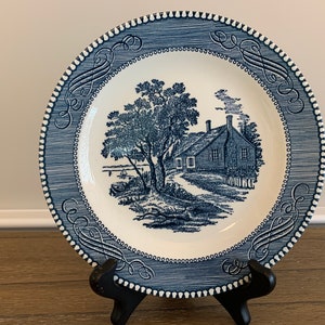 May include: Blue and white ceramic plate with a blue and white illustration of a house and tree. The plate has a blue and white border with a decorative pattern.