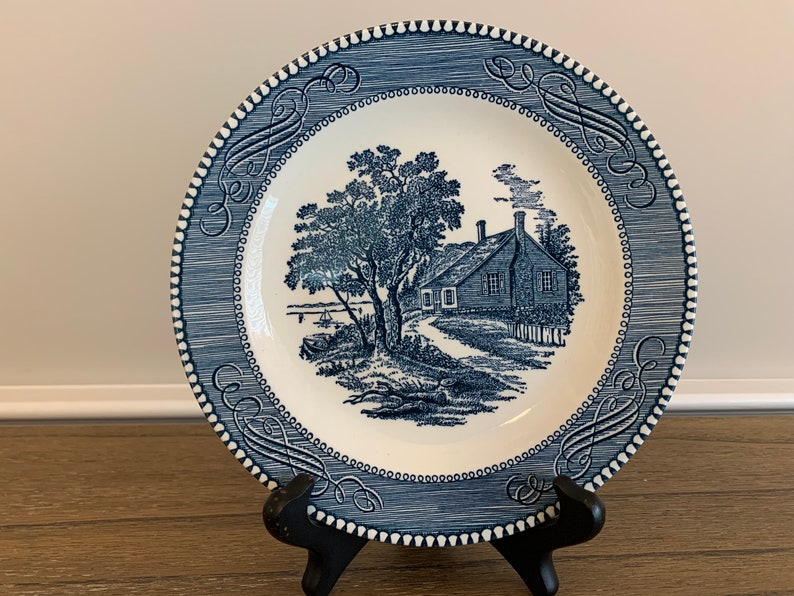 May include: Blue and white ceramic plate with a blue and white illustration of a house and tree. The plate has a blue and white border with a decorative pattern.