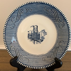 May include: Blue and white ceramic saucer with a decorative border and a steamboat design in the center.