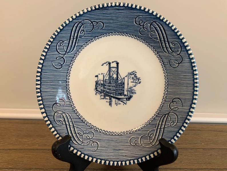 May include: Blue and white ceramic saucer with a decorative border and a steamboat design in the center.
