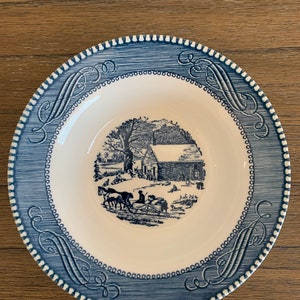May include: A blue and white ceramic plate with a winter scene of a house and people in a horse-drawn sleigh. The plate has a blue and white border with a decorative pattern.