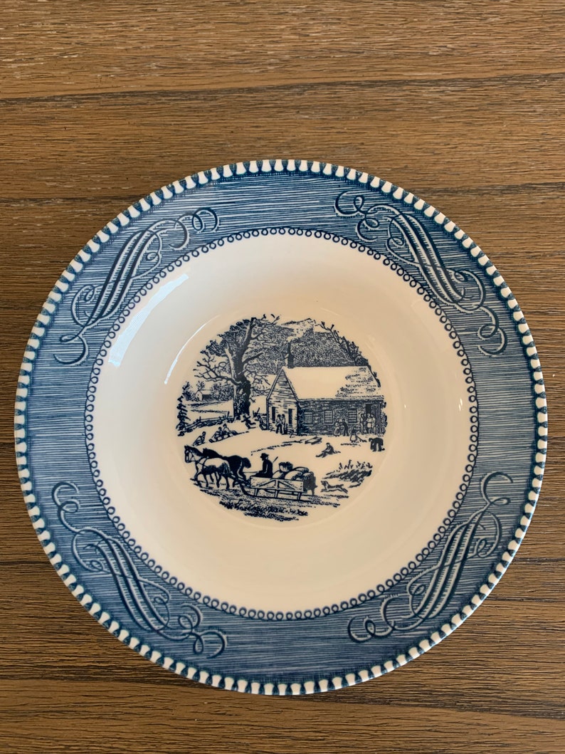 May include: A blue and white ceramic plate with a winter scene of a house and people in a horse-drawn sleigh. The plate has a blue and white border with a decorative pattern.