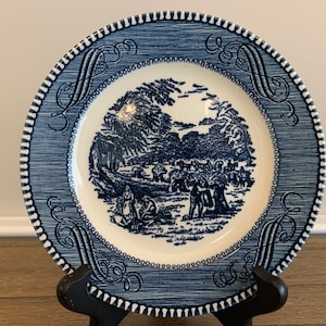 May include: Blue and white ceramic plate with a blue and white design of a pastoral scene with people and trees. The plate has a blue and white border with a scalloped edge.