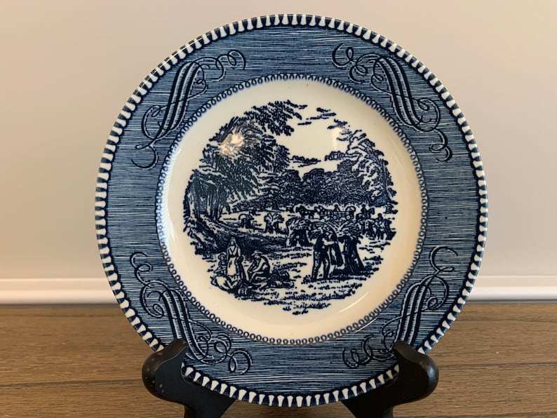 May include: Blue and white ceramic plate with a blue and white design of a pastoral scene with people and trees. The plate has a blue and white border with a scalloped edge.