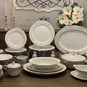May include: A complete set of white fine china dinnerware with a delicate floral pattern and gold trim. The set includes plates, bowls, cups, saucers, a serving platter, a gravy boat, and a sugar bowl.