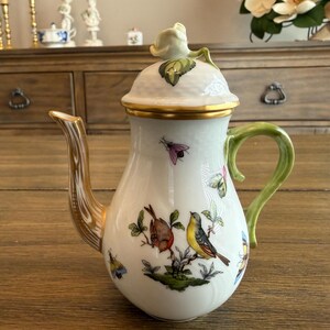 May include: A white porcelain teapot featuring hand-painted colorful birds, insects, and floral motifs. The teapot has a gold-trimmed spout, a green handle, and a lid topped with a sculpted white rosebud finial. The body has a subtle textured finish.