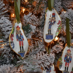 Pode incluir: Enfeites de árvore de Natal em forma de concha, cada um com um soldado quebra-nozes pintado com um uniforme colorido. Os enfeites são adornados com ouro e pendurados em fitas verdes, em um fundo de pinheiro nevado.