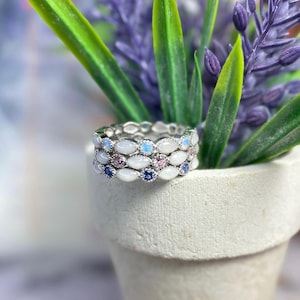 May include: Stackable silver rings with white, blue, and pink gemstones. The rings are set in a delicate, repeating pattern. The rings are displayed in a small white pot with green and purple plants.