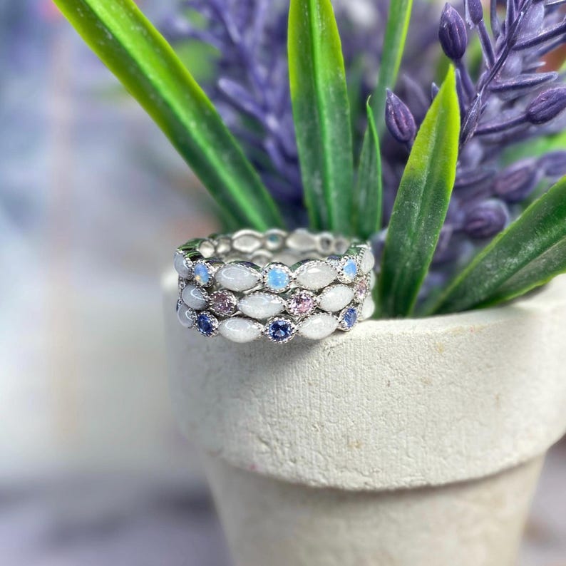 May include: Stackable silver rings with white, blue, and pink gemstones. The rings are set in a delicate, repeating pattern. The rings are displayed in a small white pot with green and purple plants.
