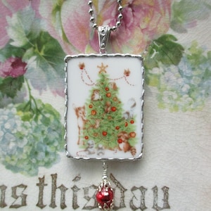 May include: A silver chain necklace with a square porcelain pendant featuring a vintage Christmas tree illustration with red ornaments and small animals. A red glass bead dangles from the pendant.